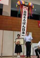 高校そろばん日本一のくす玉の下で賞状を掲げる西奈瑠美さんの写真