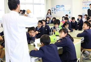 理科室で前に立つ白衣を着た男性を見つめる生徒たちの写真