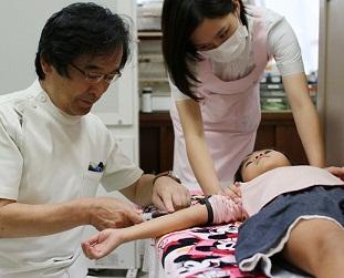 診察台の上で腕を伸ばした女の子に注射をする医師の写真