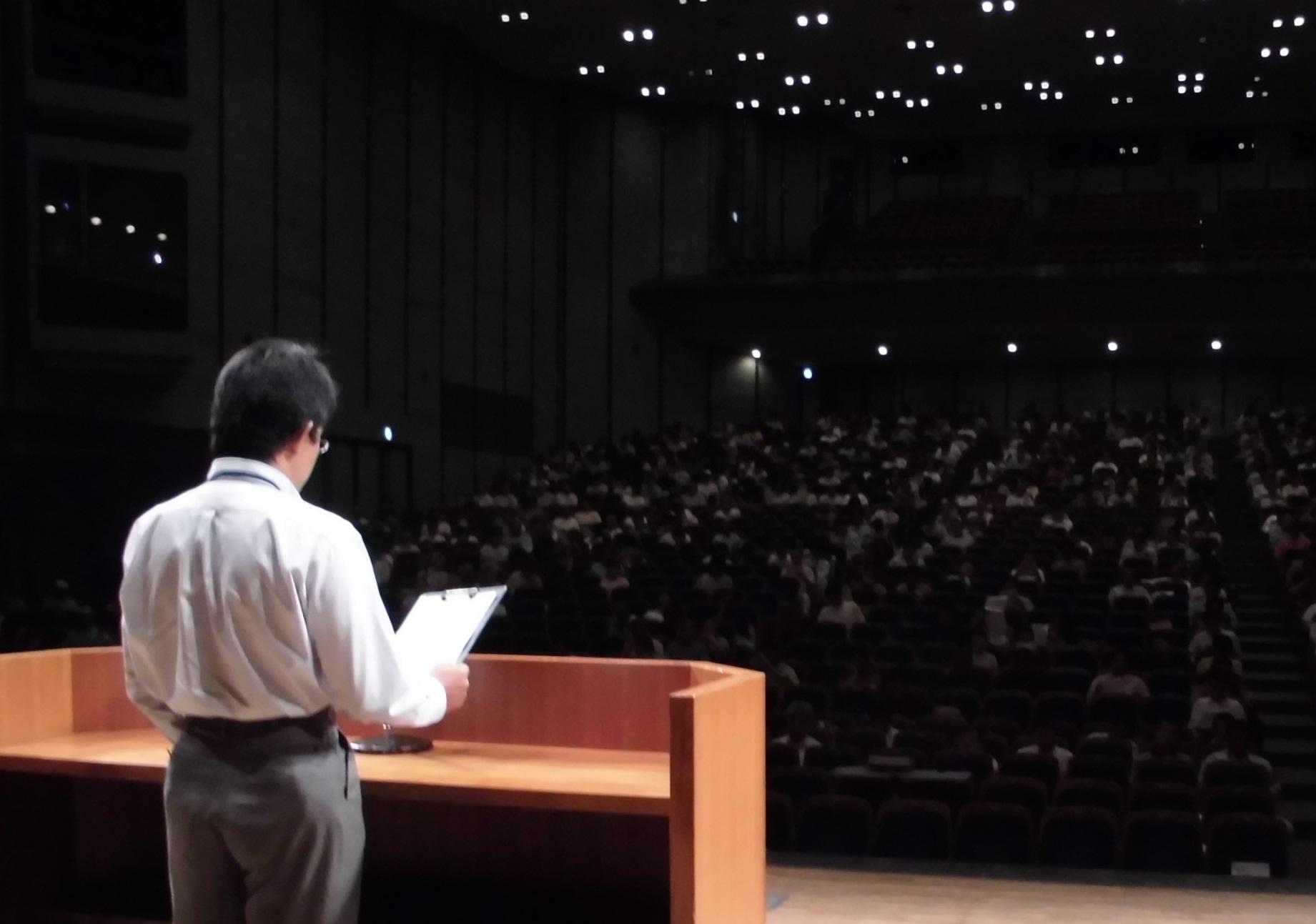 受講者たちの前で男性の講師がステージ上に立って講習を行っている様子を撮影した写真