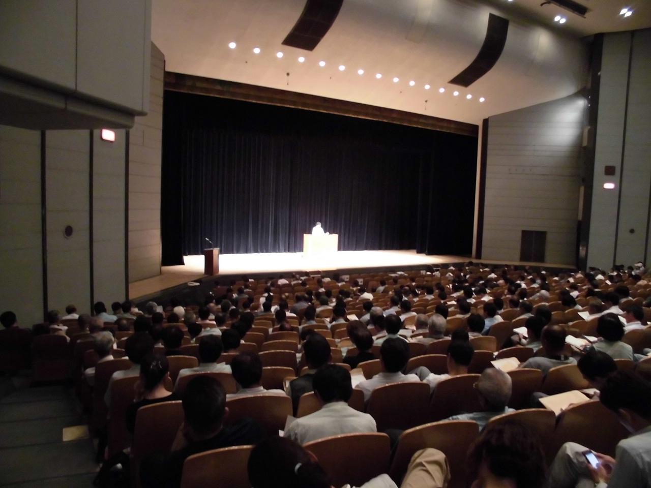 男性の講師がステージ上に立って講習を行っている様子を、受講者席側から撮影した写真