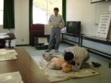 男性が心肺蘇生人形を使って救命訓練をしていて近くで別の男性が見守っている写真