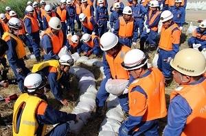 白いヘルメットを被りオレンジ色や黄色のベストを着た多くの作業員たちが土のうを三日月型に繋げている写真