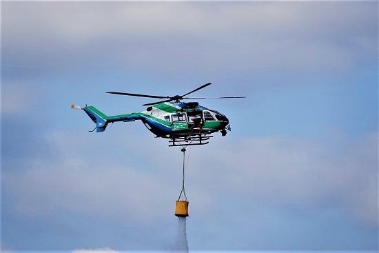 緑色と青色のヘリコプターから黄色いものが下がり、消火活動を行っている写真