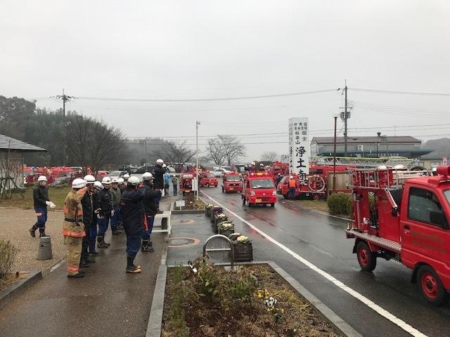 雨の中赤い小さな消防車が車両行進している様子と歩道に並んでいる消防団員たちの写真
