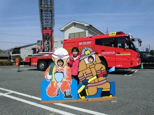 はしごを立てている消防車の前で赤い服を着てVサインをしながら記念撮影をする女の子の写真