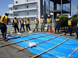 ブルーシートの上にパイプと土のうが置かれていて消防隊員と作業員が眺めている写真