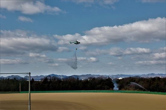 ヘリコプターが平地の上空を飛び、消火訓練を行っている写真