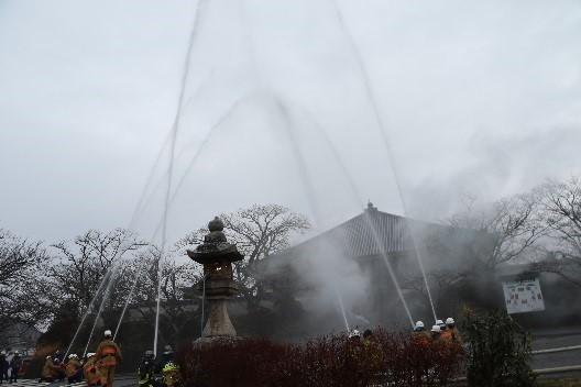 寺院の上空めがけて消防団員が構えたそれぞれのホースから一斉に放水している写真