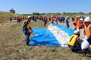 草原にブルーシートを敷いて多くの土のうを積んでいるオレンジ色や黄色のベストを着た多くの作業員たちの写真