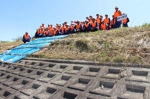 土手にブルーシートが敷かれ、土手の上にオレンジ色のベストを着た作業員たちが並んで立っている写真