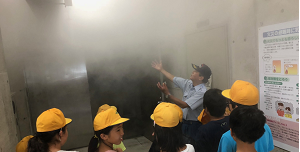 煙が充満した部屋の前で職員の説明を聞いている生徒たちの写真