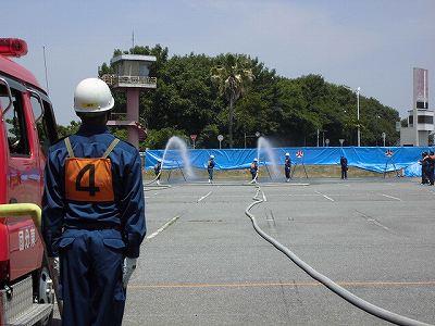 消防車の脇に消防隊員が立っていて奥で放水をしている写真