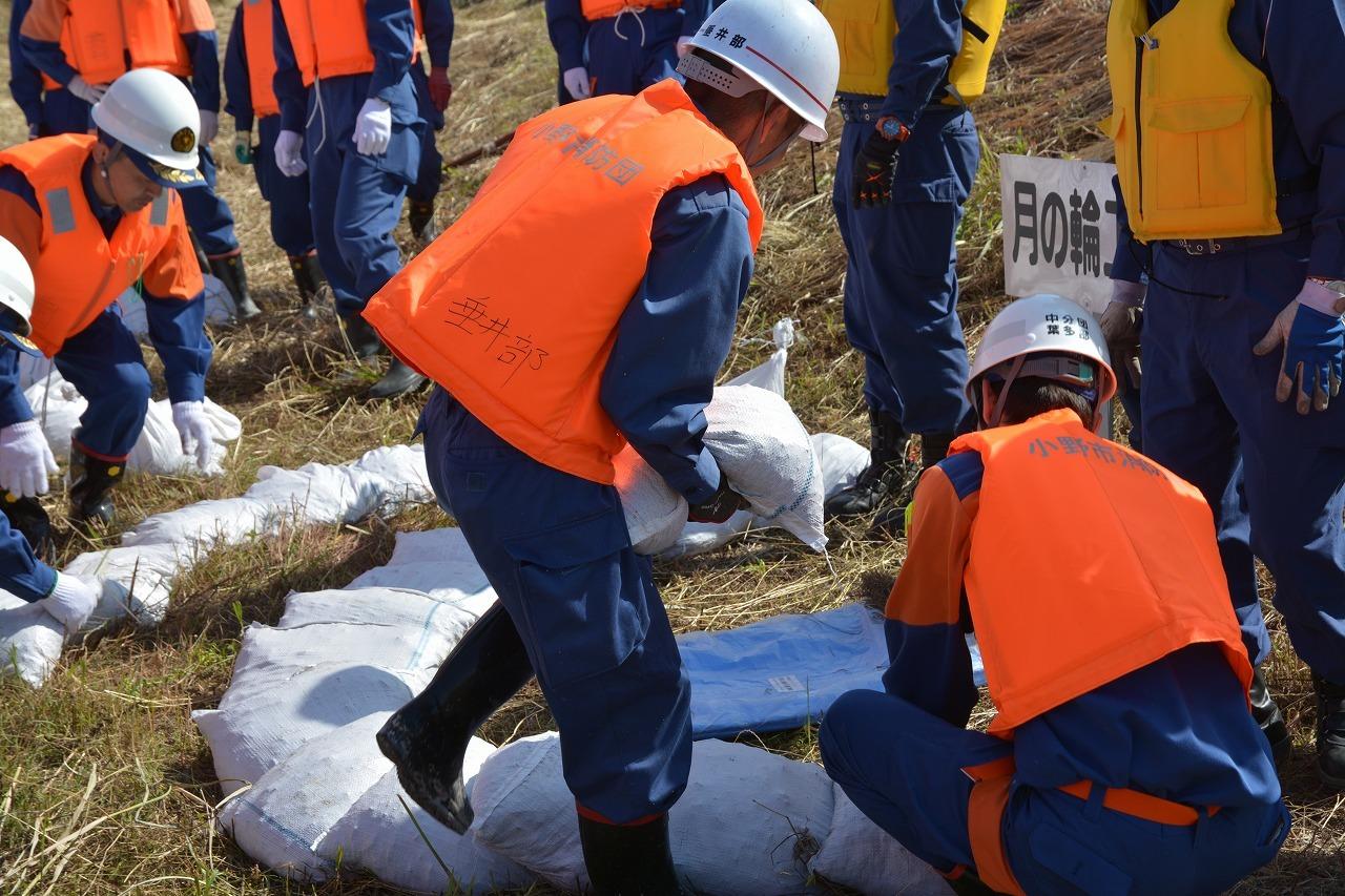 オレンジ色のベストを着た消防関係者の人々が三日月型に土のうを積んでいる写真