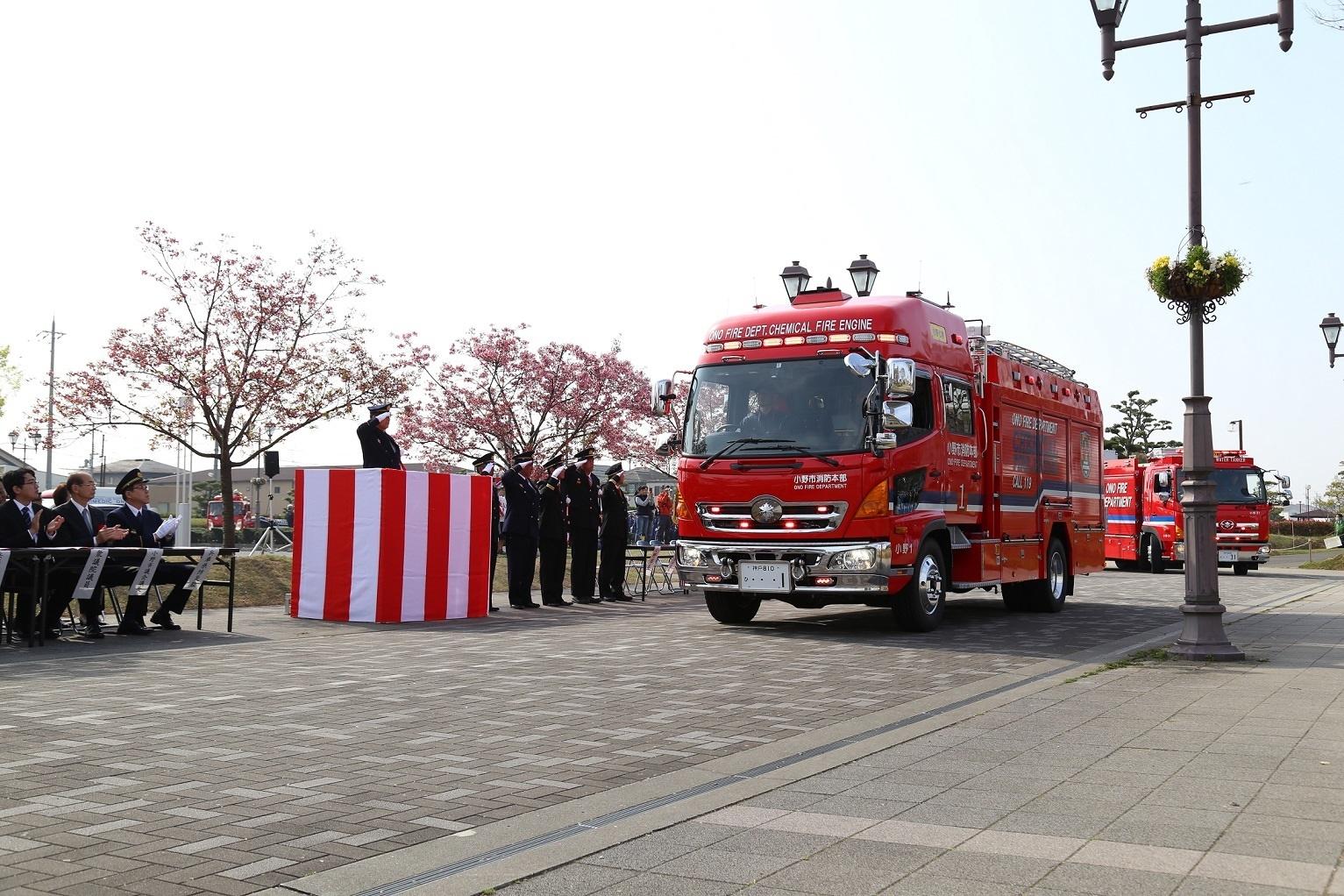 制服を着た人々が紅白の幕の前に立ち、そこに消防車が停まっている写真