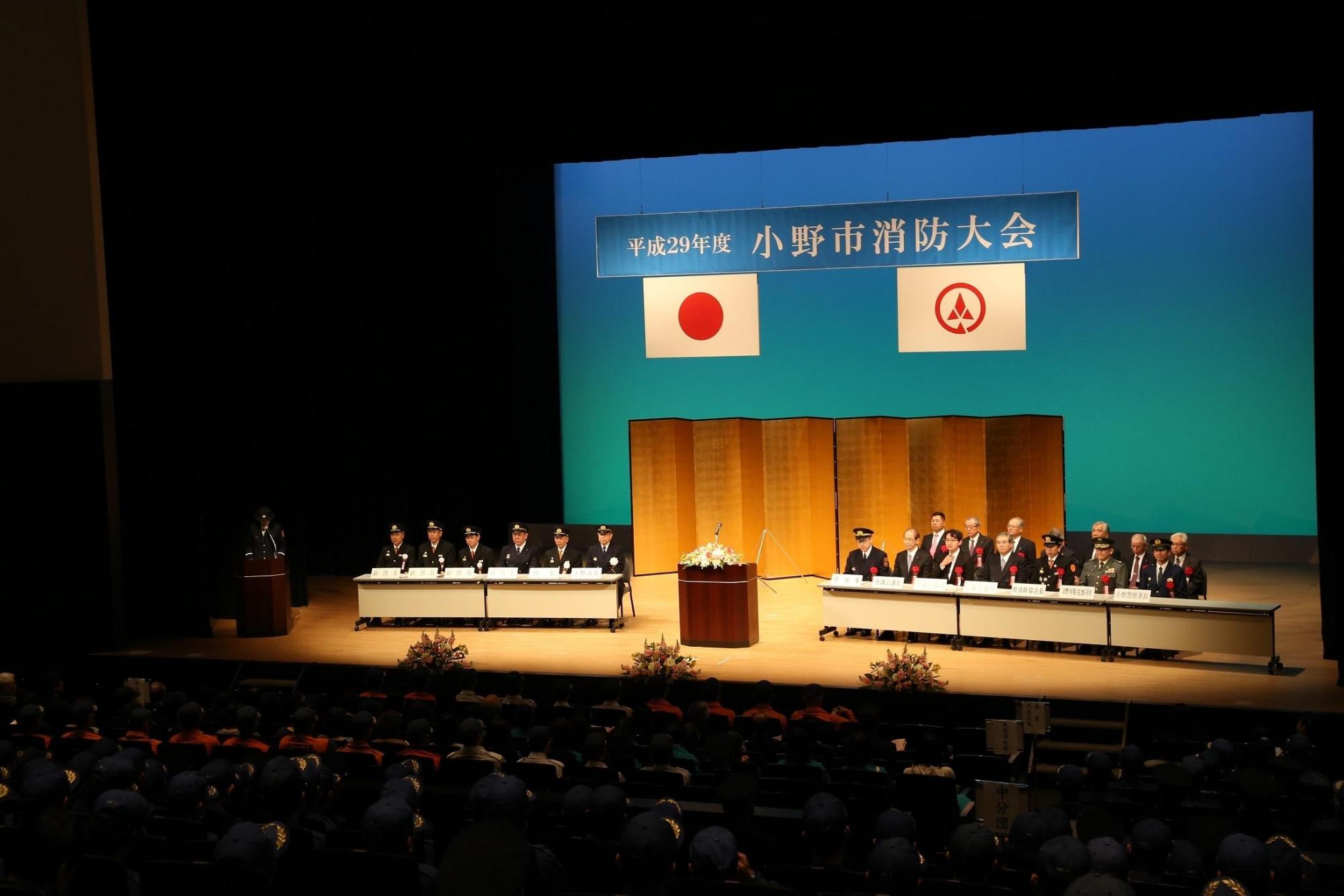 小野市消防大会と書かれた幕をバックにして人々が腰掛けている写真