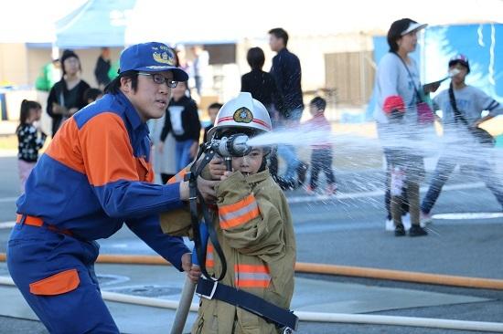 小さな男の子が消防隊員と一緒に放水体験をしている写真