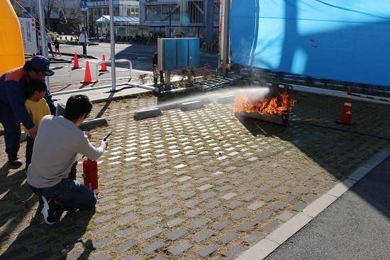 白い上着を着た男性が消防隊員や男の子と一緒に消火器を持って初期消火体験をしている写真