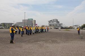 オレンジ色や黄色のベストを着た大勢の作業員たちが黄色のベストを着た作業員の男性と向き合っている写真