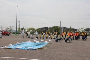 オレンジ色や黄色のベストを着た作業員たちが並んでいる前にブルーシートが敷かれ土嚢が置かれている写真
