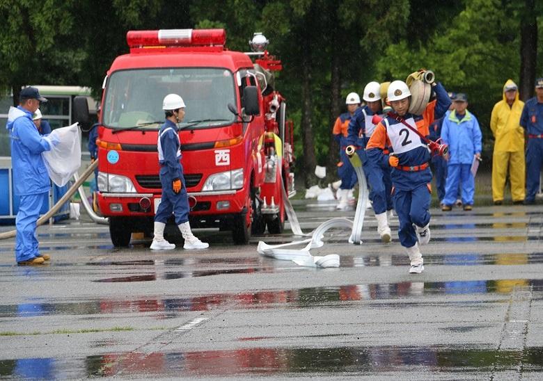 消防車の右側を多数の消防隊員が手前に向かって巻かれた送水ホースを背負って走っている写真