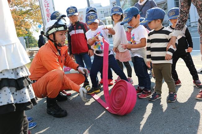 実際に使う放水ホースを手にとって興味津々に見ている子供たちとサポートする隊員の写真