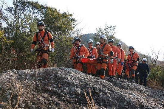山岳を赤い担架を持って移動するオレンジ色の防護服を着た隊員たちの写真