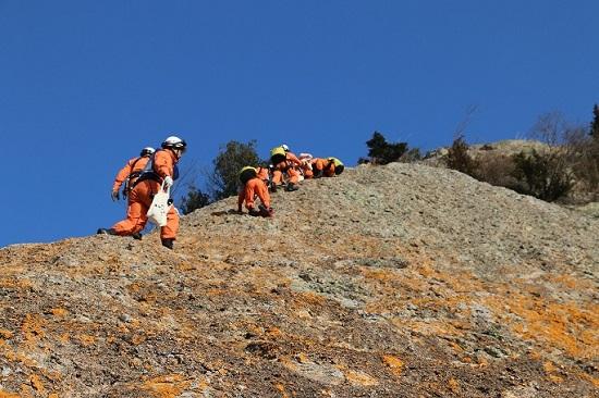 急な角度の山岳を登るオレンジ色の防護服の隊員たちの写真