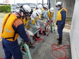 ヘルメットを被った消防隊員と作業員がバリケードのパイプを使って結索訓練をしている写真