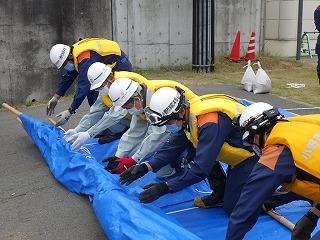 黄色いベストを着て白いヘルメットを着た作業員たちが並んでブルーシートを敷いている写真