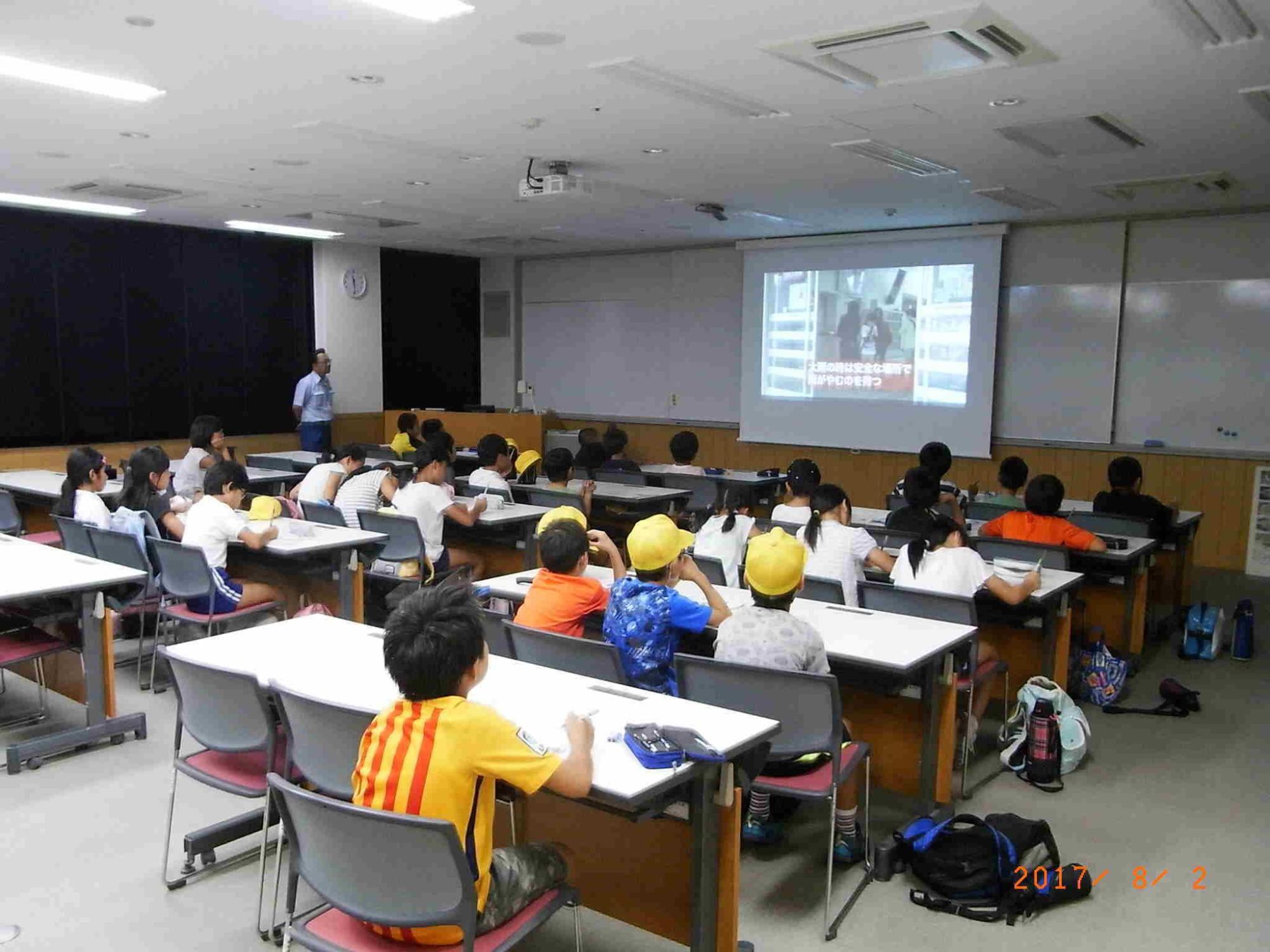 白い机が並び生徒がそれぞれ着席しスクリーンの映像見ているところを右後から撮った写真
