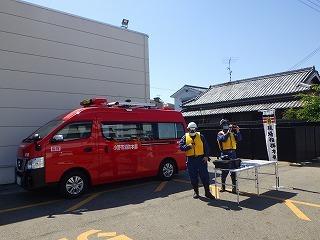 赤い車の傍に黄色のベストを着た作業員2人が立っている写真