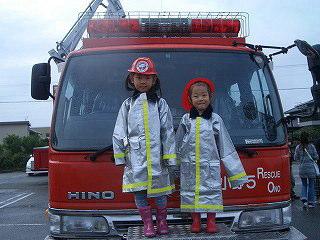 消防車の前で防護服を着て記念撮影している子ども二人の写真