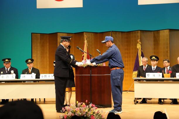 青い制服を着た男性が黒い制服を着た男性から表彰状を受け取っている写真