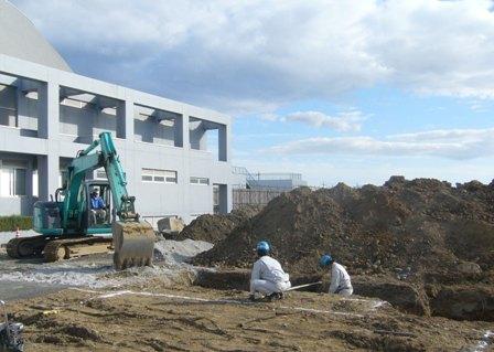 盛り土と溝があり溝で作業員2人が計量を行い左側にショベルカーが停車している写真