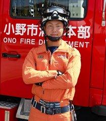 赤い消防車のバックに、ゴーグルのついたヘルメットをかぶり、オレンジ色の消防士の制服を着て腕を組んでいる様子の吉田 圭祐さんの写真