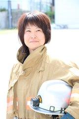 ベージュ色の消防士の制服を着て、銀色のヘルメットを左腕で抱えるように持って写真に写る田中 利奈さんの写真