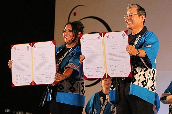 リンゼイ市女性市長と小野市男性市長がハッピを着て、調印式で結んだ調印書を並んで前に掲げている写真